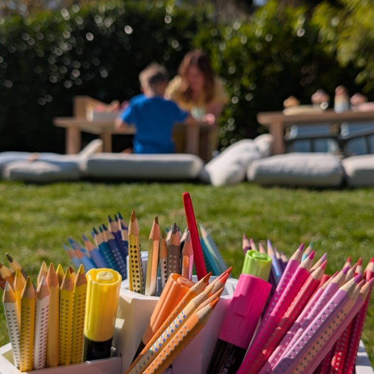 Kinderbetreuung im Freien Bunte Stifte in einem Behälter, mit Kindern und einer Frau im Hintergrund beim Spielen.